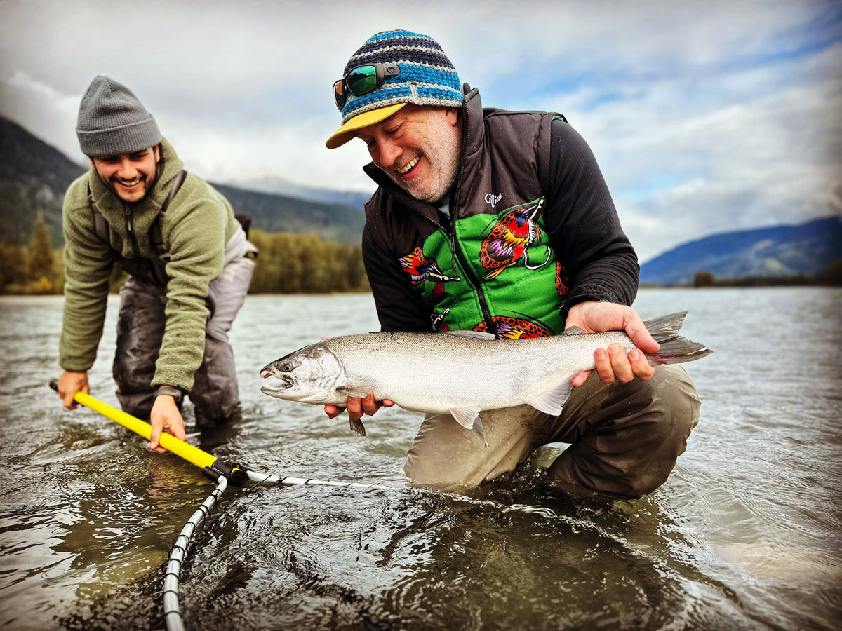 A man in a colorful beanie and green chest waders holds a large salmon in shallow water while another person uses a yellow net to guide it back into the river. Mountains and a cloudy sky are visible in the background.